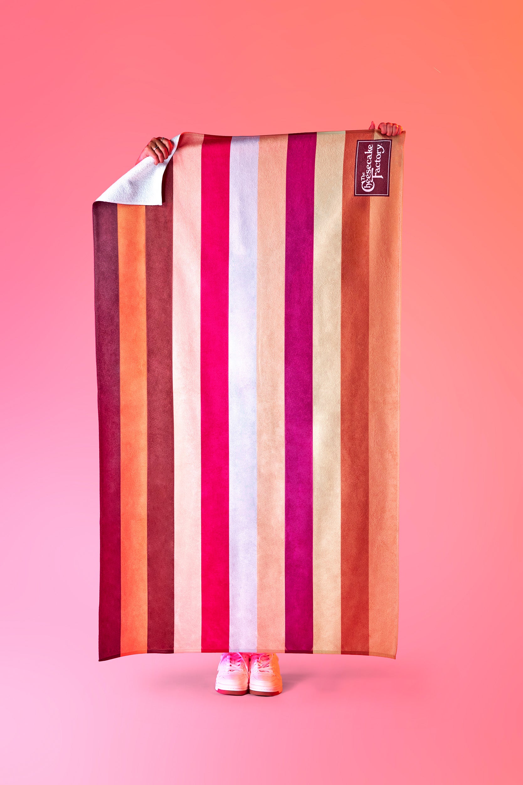 Full view of a beach towel printed with The Cheesecake Factory's signature stripes across the towel horizontally and brown boxed logo in the bottom right. 