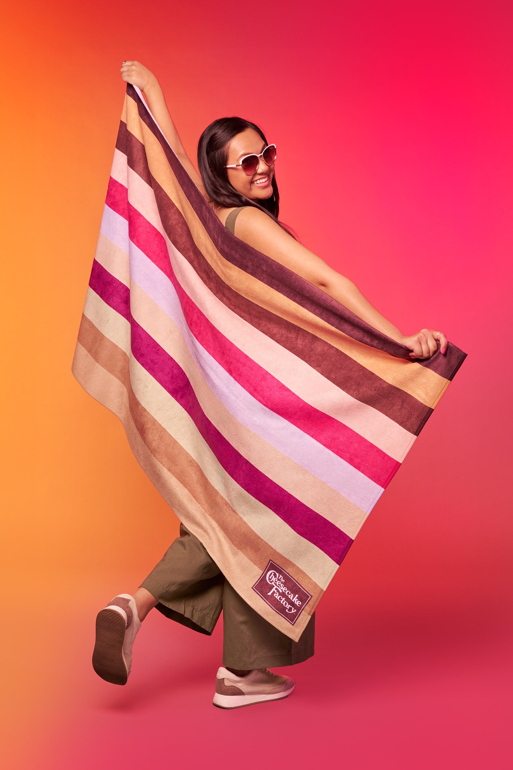 Woman holding a beach towel printed with The Cheesecake Factory's signature stripes across the towel horizontally and brown boxed logo in the bottom right. 